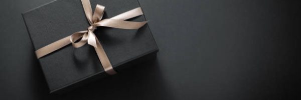 One gift wrapped in dark black paper with luxury bow on dark background. Horizontal with copy space.